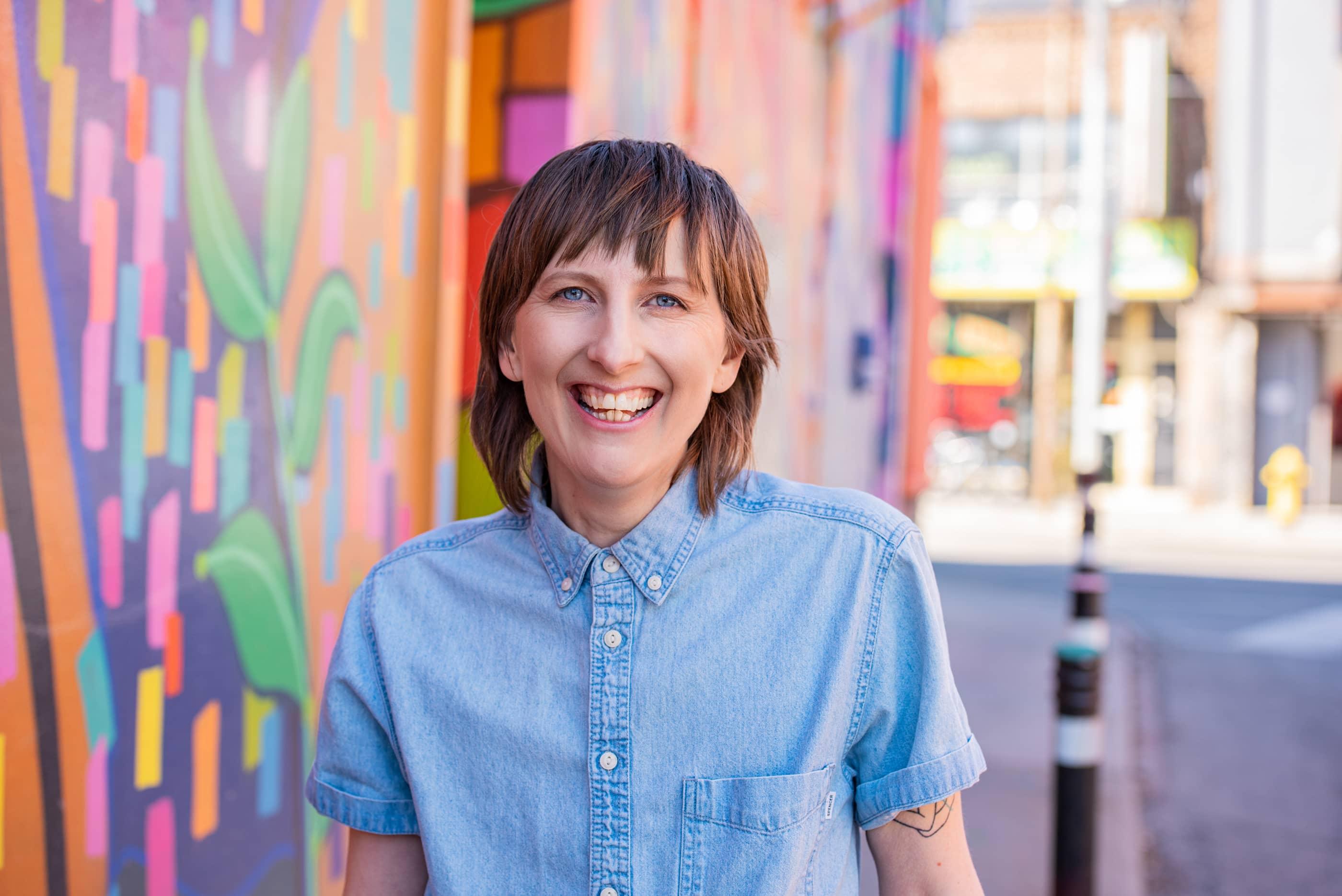 Nicole wearing a blue button down shirt smiling on a street with a colourful mural in the background